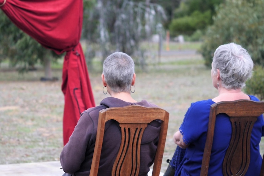A view from the behind of two women sitting on chairs outside with red curtains visible in front of them.