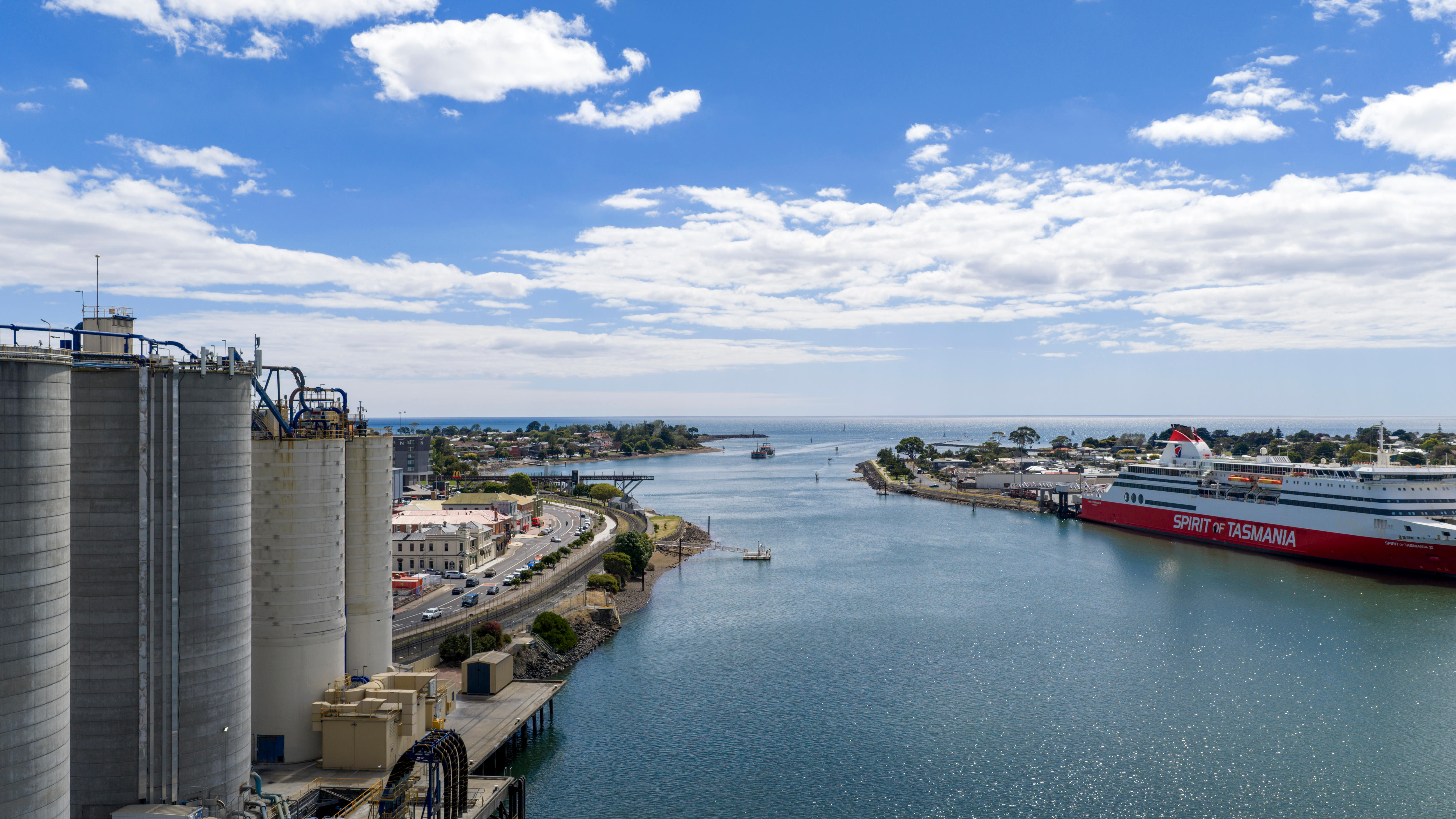 An aerial view of a calm ocean port, with industrial silos on the left bank and a large passenger ferry berthed at right.