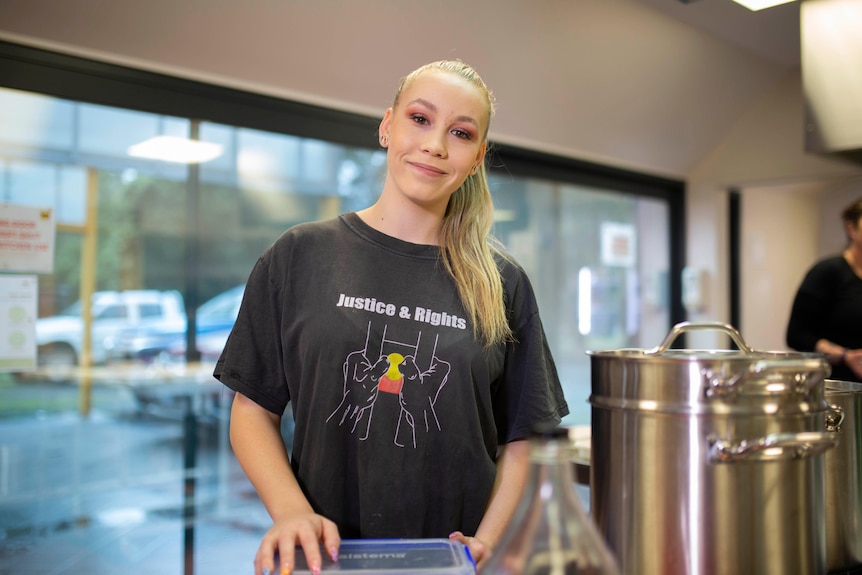 A woman in a kitchen smiles at the camera, she wears an Aboriginal justice shirt