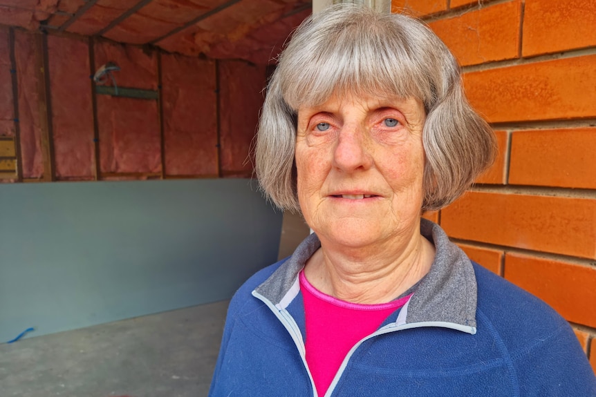 A woman with grey hair stands in front of a brick wall.