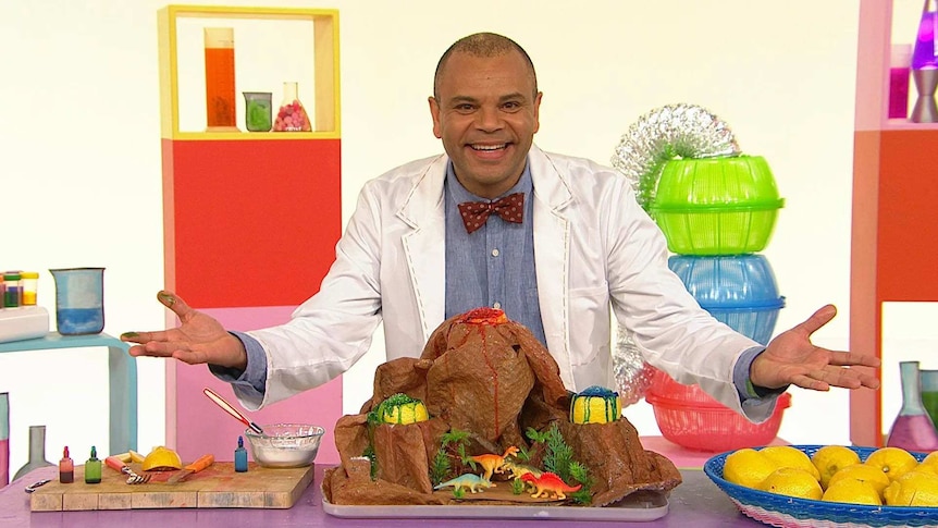 Luke on the Play School Science Time set wearing a lab coat standing behind a craft volcano with toy dinosaurs