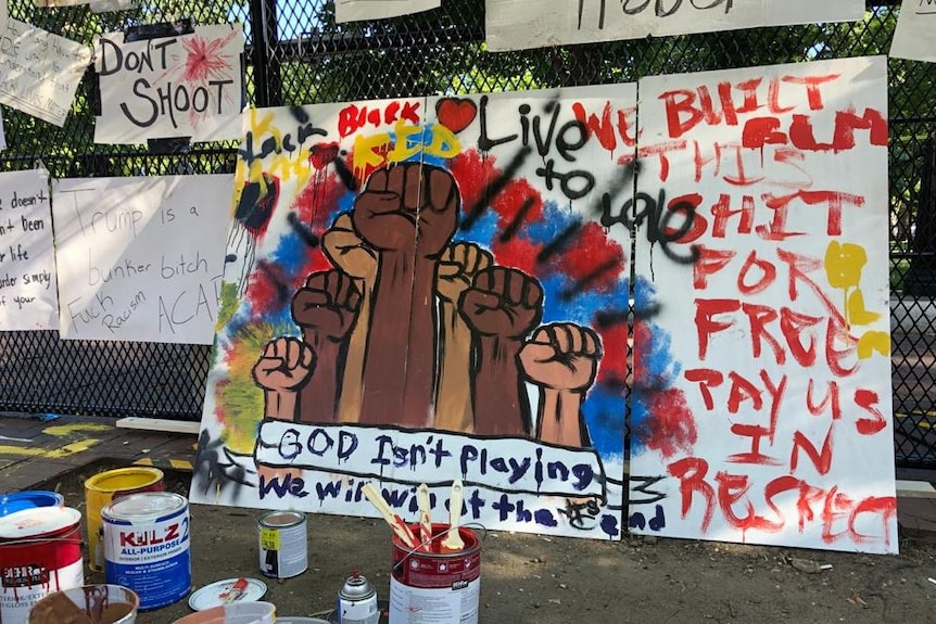 Paint cans and brushes sit on the ground besides signs against a fence reading Black Lives Matter.