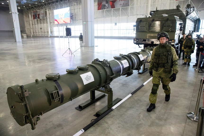 Военный офицер, одетый в зеленое, идет горизонтально через сарай мимо большой крылатой ракеты. 