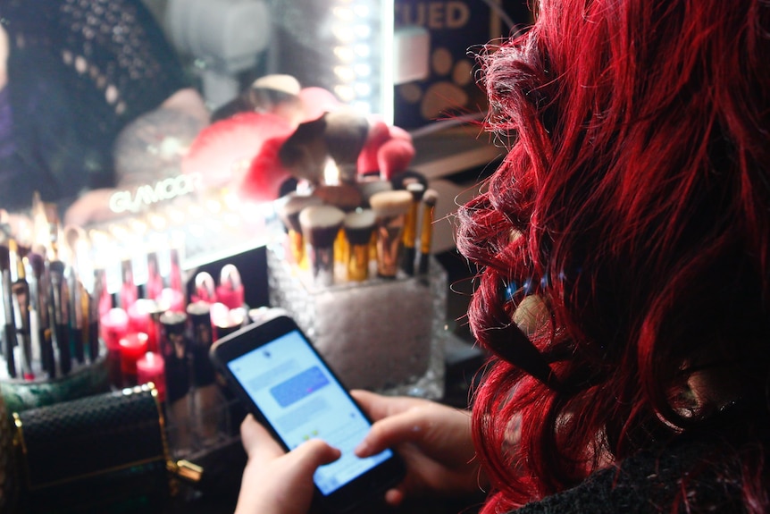 A person with long hair sits in front of a table with makeup on it, using their hands to send a message on their phone.