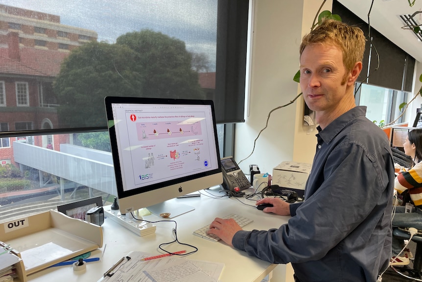 Peter wears a blue collared shirt and stands at a computer on a work desk in front of a large window