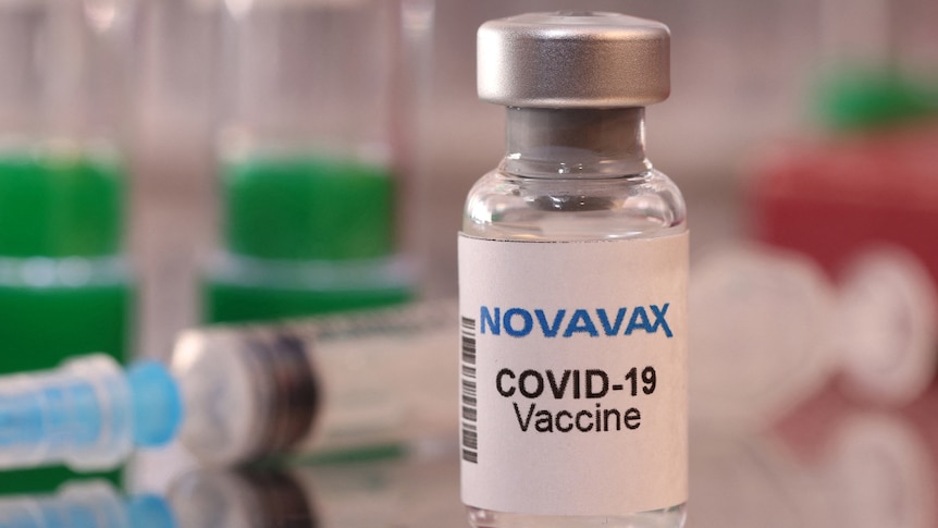 A vial of Novavax COVID-19 vaccine with a blurred needle in the background.