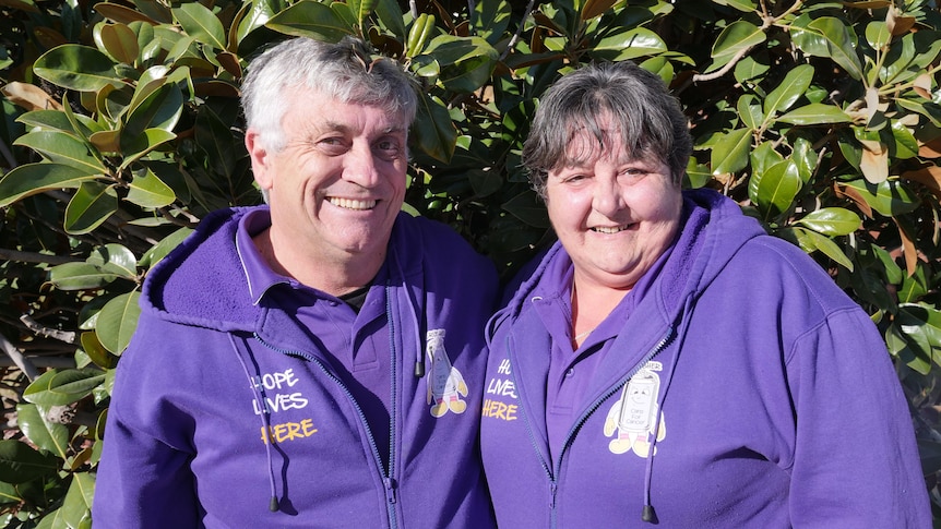 A man and a woman wearing matching hoodies stand smiling in front of some shrubbery.
