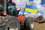 A farmer waves from a tractor that has a Ukrainian flag attached to it.