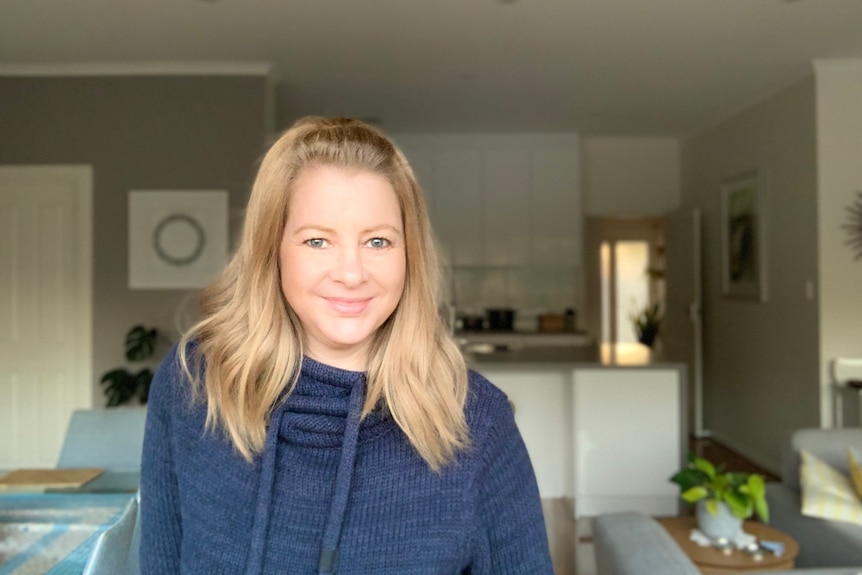 A woman with bronde hair stands in her lounge/kitchen area and smiles into the camera. She wears a navy hoodie.