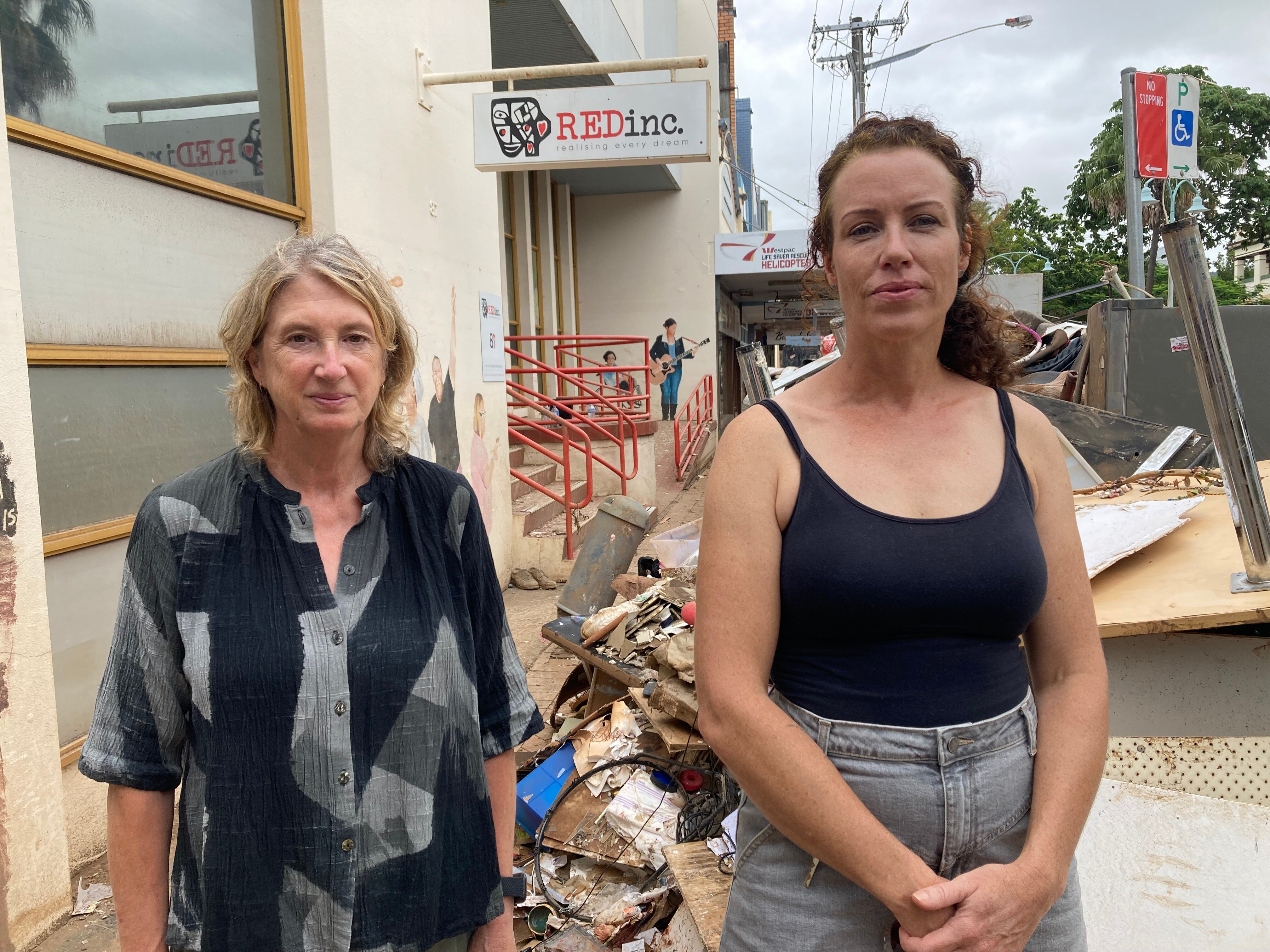 A woman in a black and grey shirt, and a woman in a black singlet, standing near a pile of rubbish outside a building.