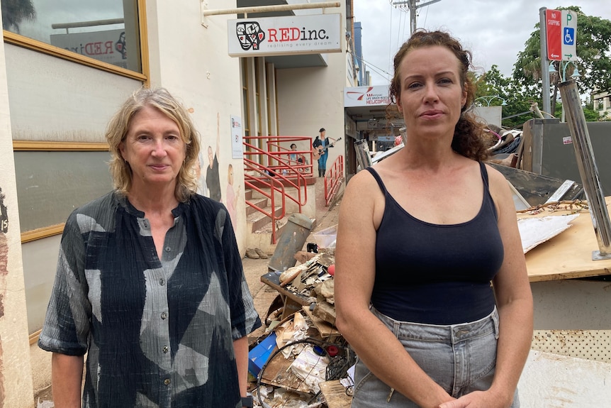 A woman in a black and grey shirt, and a woman in a black singlet, standing near a pile of rubbish outside a building.