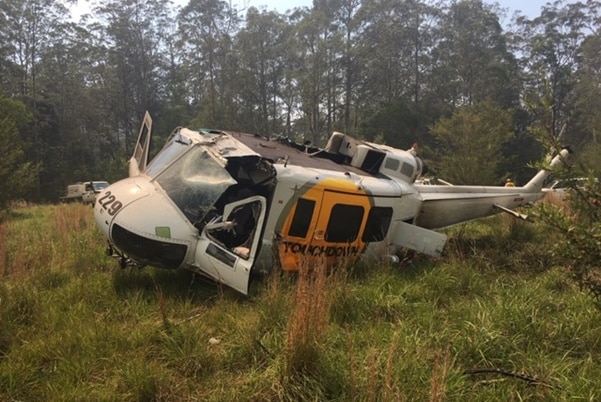 A helicopter tilts on its side, with no rotors, in a clearing surrounded by trees.