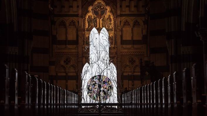 An illuminated artwork below a church alter, sitting in front of rows of pews.