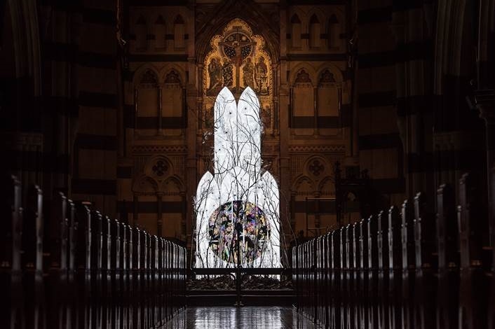 An illuminated artwork below a church alter, sitting in front of rows of pews.