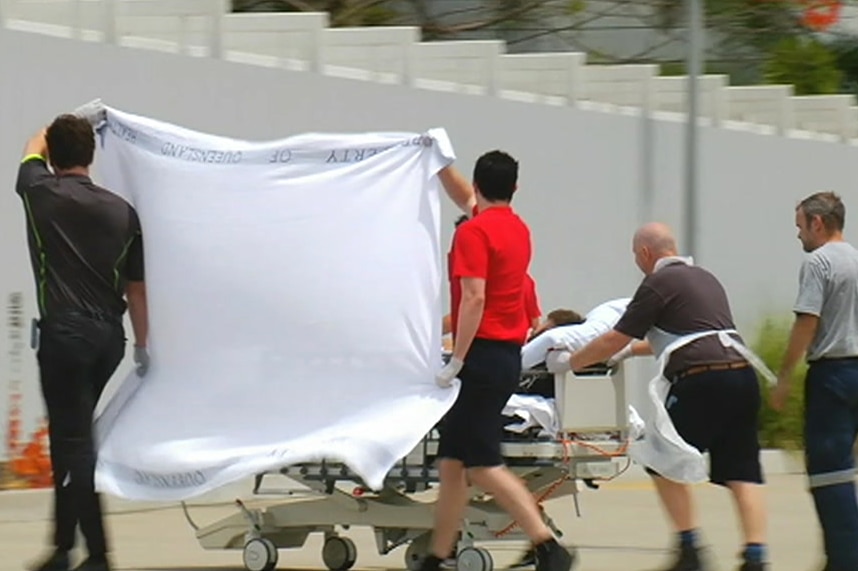 A man is wheeled on a gurney by hospital staff. Two men hold up a sheet to block the view of the media.