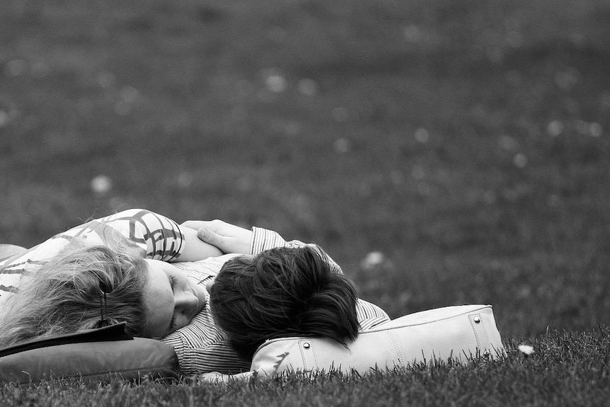 A couple lie in the grass, heads tilted towards each other.