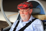 A smiling gentleman sitting in a glider wearing a tartan beret.