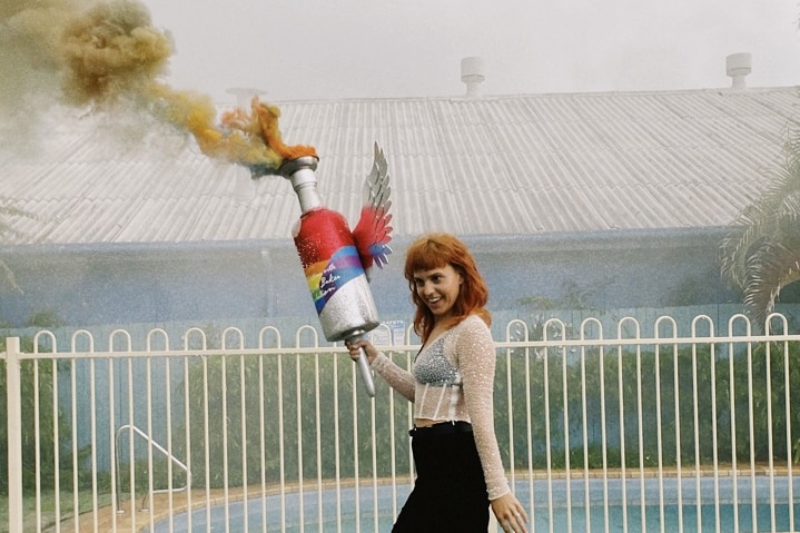 A woman with red hair holds a large bottle releasing rainbow smoke. She wears black pants and silver shoes.