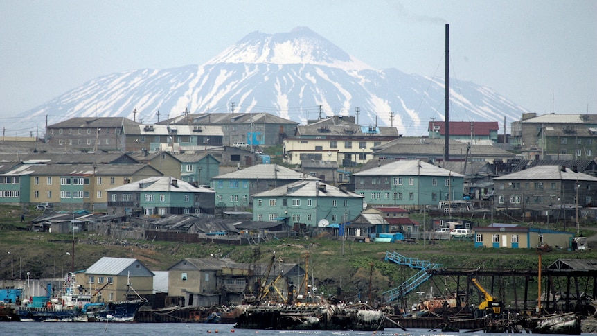 A little fishing village of blue wooden shacks on the water's edge, with a snow capped mountain in the background