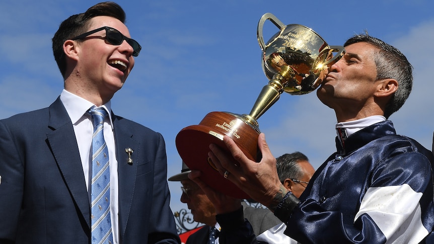 Young trainer laughs as Jockey kisses Melbourne Cup trophy.