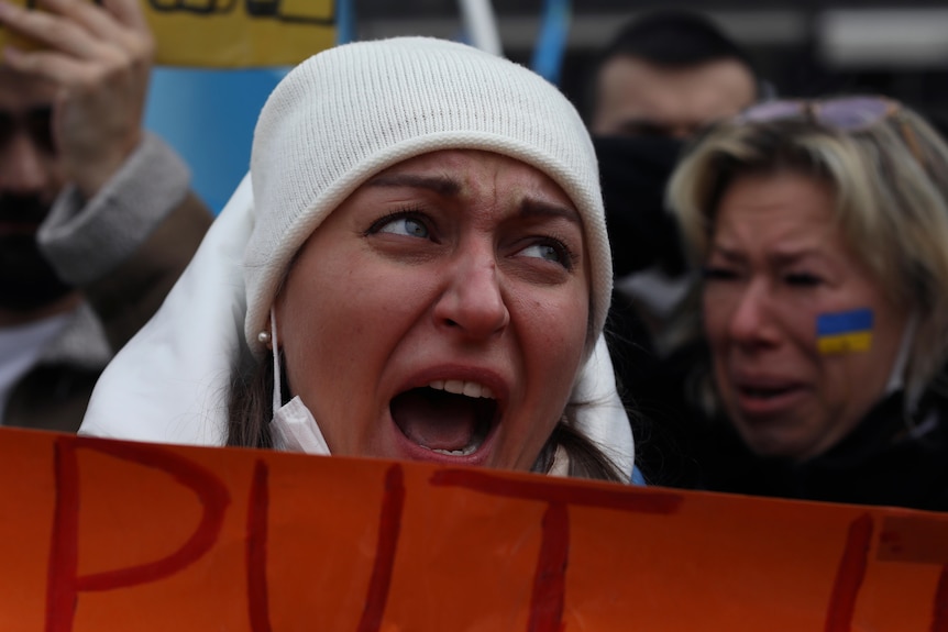 A woman wearing a white beanie and face mask around her chin screams with her mouth wide open, while holding a red sign.