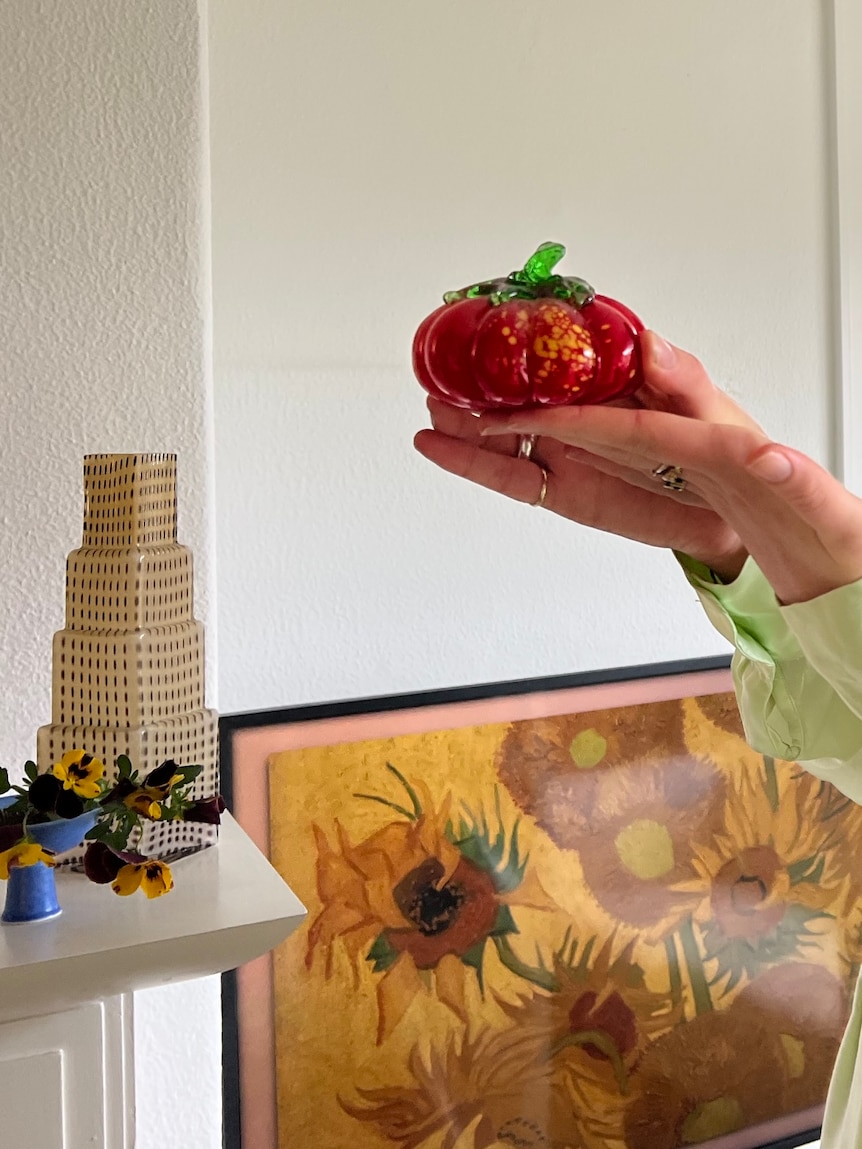 Hattie holds a red glass heirloom tomato, with a white glass skyscraper building vase on the mantelpiece behind.