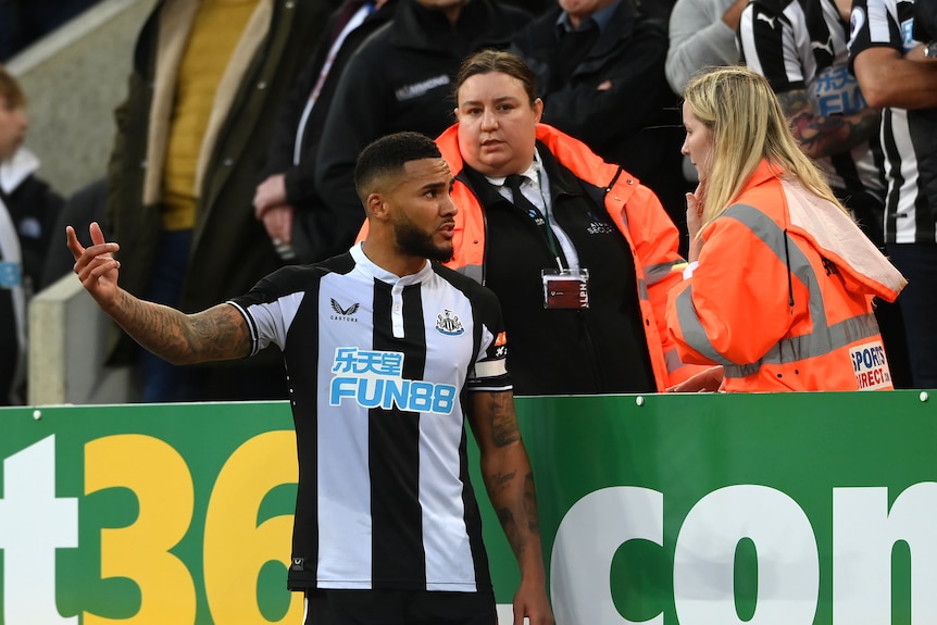 Jamaal Lascelles looks concerned and gestures as he speaks to a woman wearing a high-vis jacket on the side of the pitch