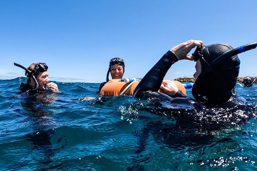 A woman in a wetsuit laughs as she speaks to divers on the surface in the open ocean.