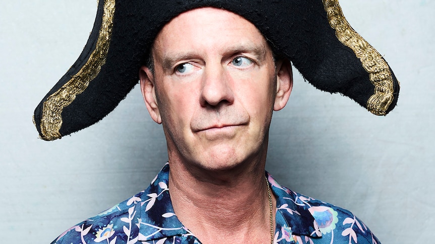 Fatboy Slim wears a large old style sailor hat. He looks disinterested and wears a brightly coloured party shirt.