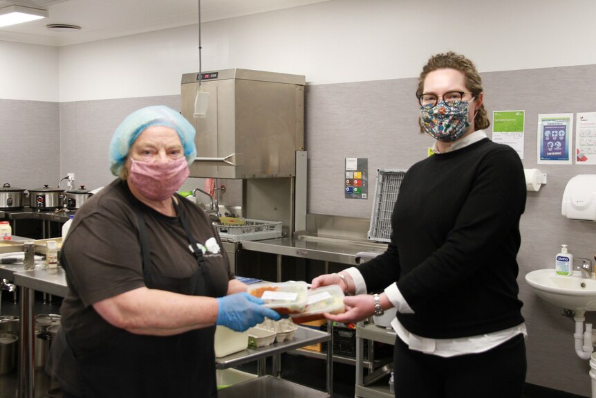 One woman wearing a hair net and mask passes a pre-prepared meal to another woman wearing a mask.