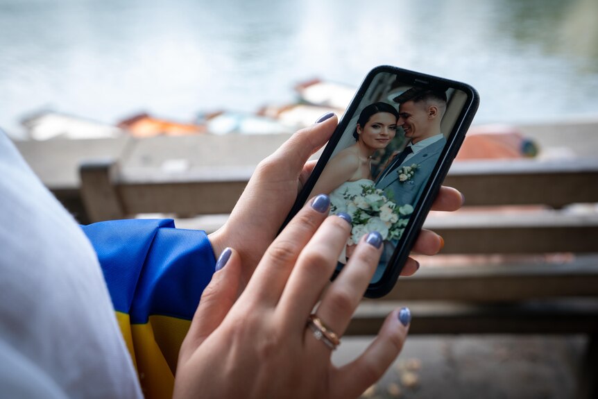 Женщина с серо-голубым лаком на ногтях и двумя кольцами стучит по экрану телефона, на котором изображена свадебная фотография.