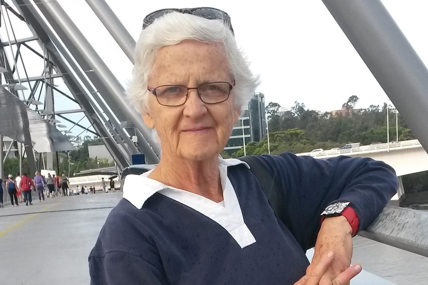 Older woman, slightly smiling, grey hair, sunglasses on top, wears glasses, blue shirt white collar. Stands in front of bridge.