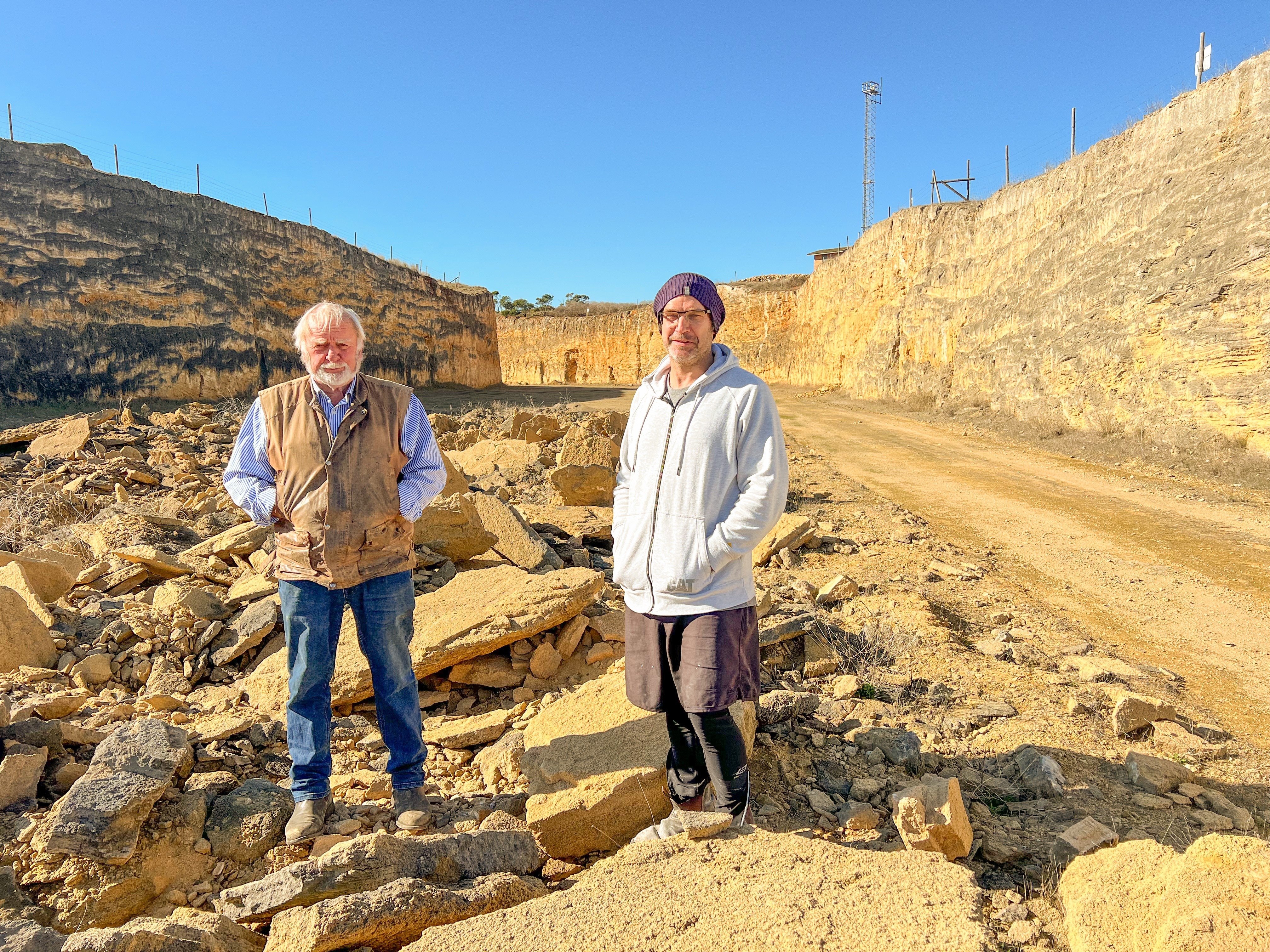 Two men in farm clothes stand on loose rock in a sandstone quarry