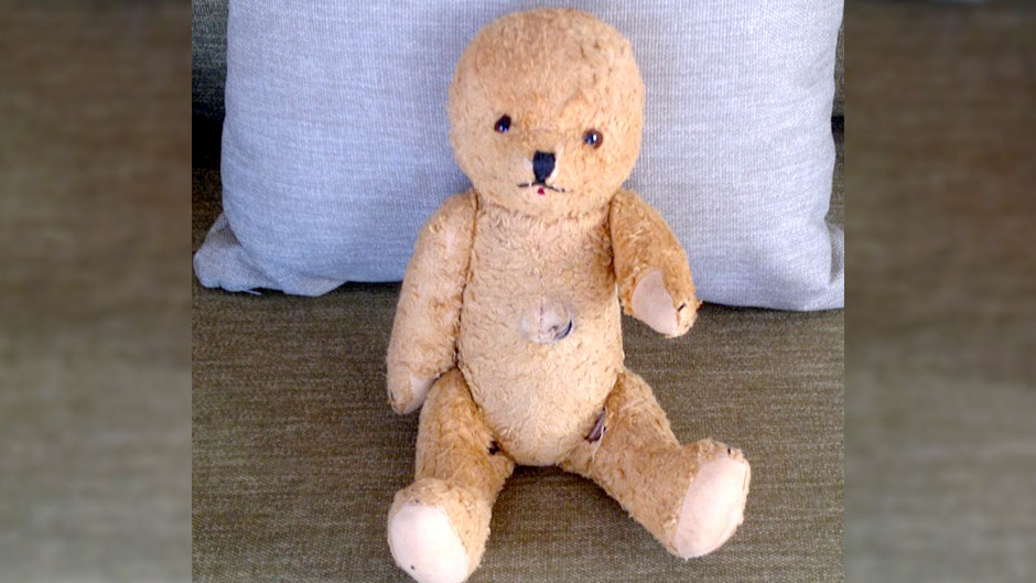 A worn teddy bear with no ears sits propped against a pillow.
