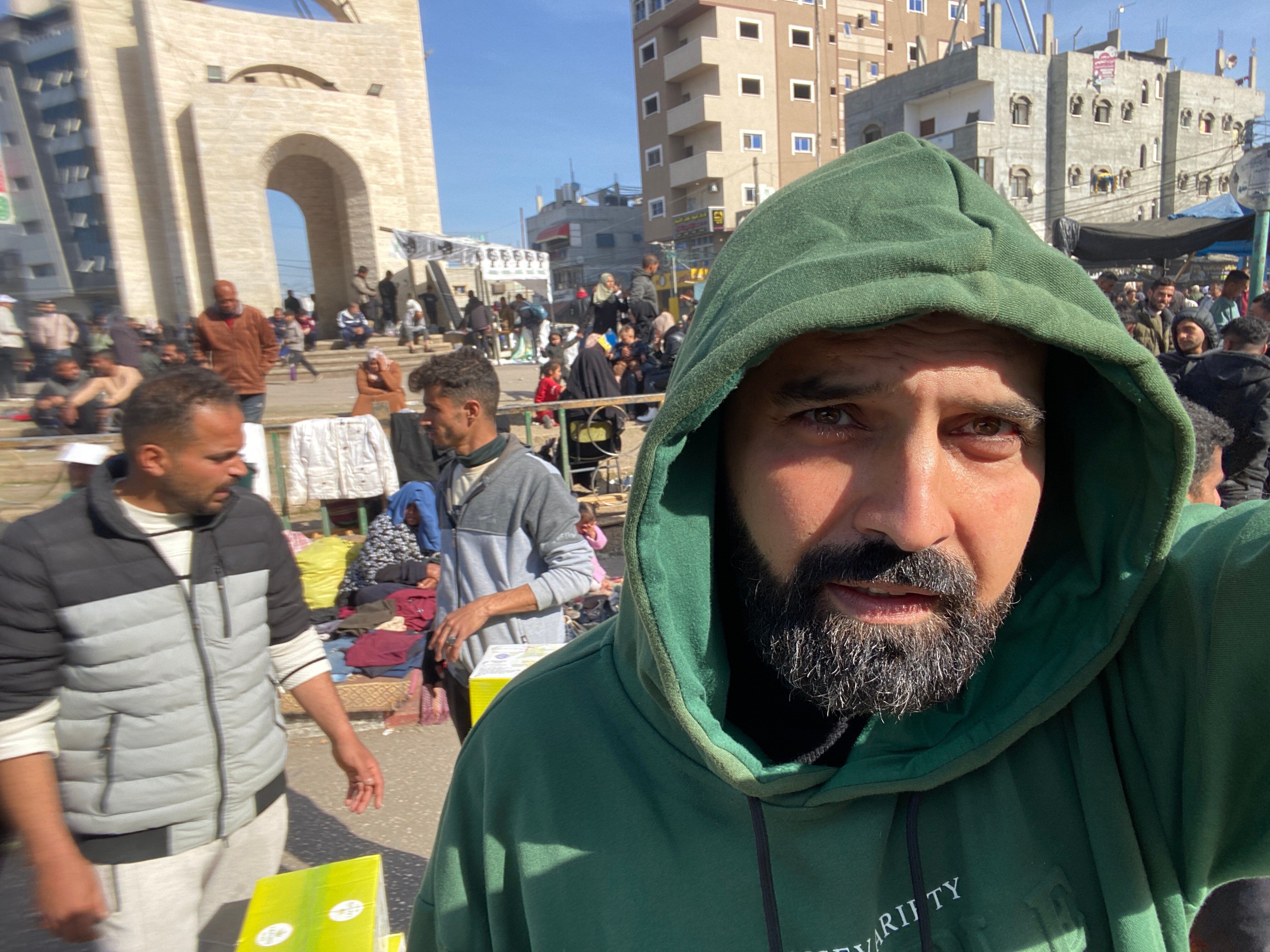 A man stands in a crowded street wearing a green hoodie. 