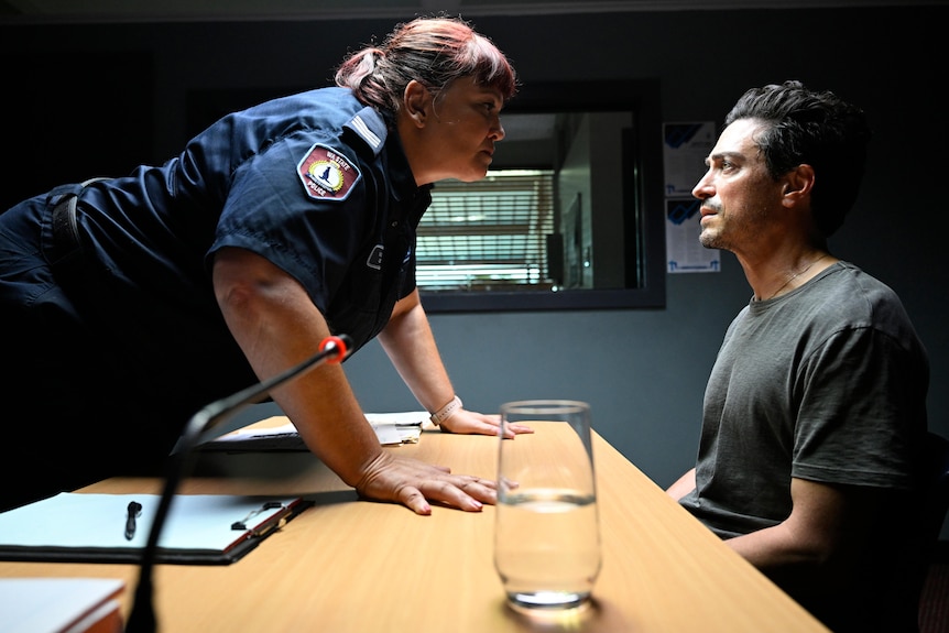 A man sits in a police station as a female police officer stares him down.