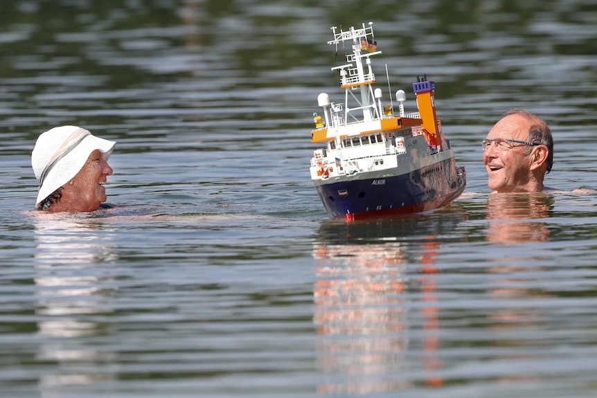 A couple swims beside a model making boat in a lake in Ertingen, Germany, Wednesday, June 26, 2019.