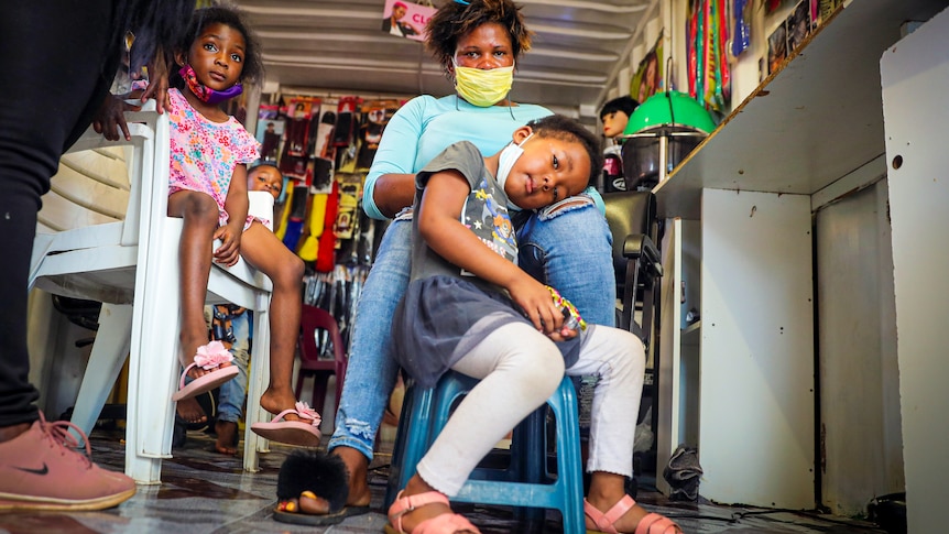 A woman in a blue face mask sits with a small girl on her lap, while another girl sits on a chair next to her