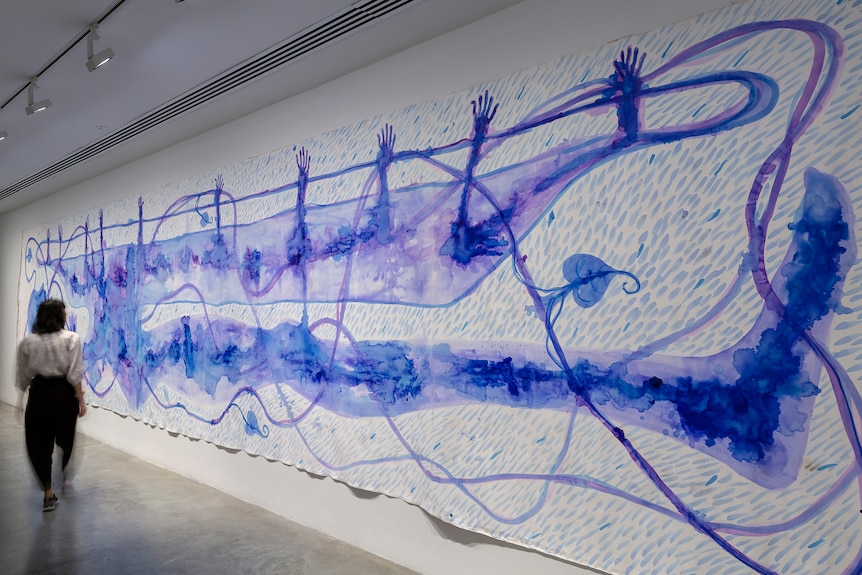 A long watercolor painting takes up much of a gallery wall, with blue hands sticking out of a body of water