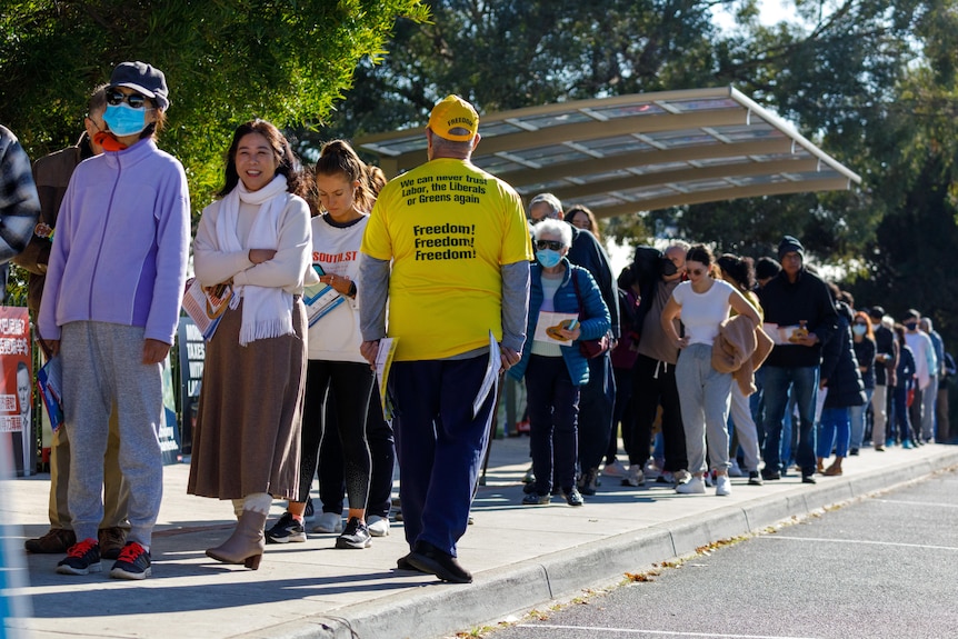 A United Australia Party volunteer in yellow walks past voters lining up for a polling booth.