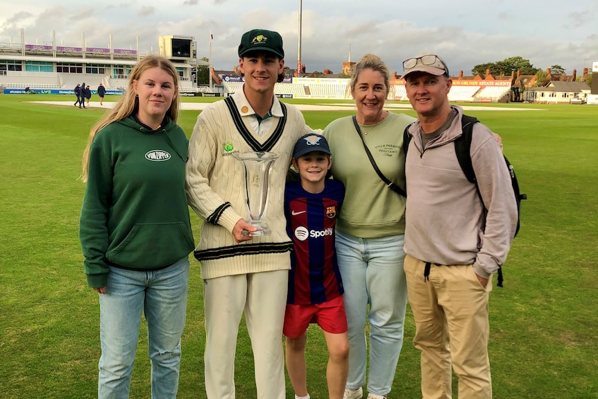 A family smiles, with one in cricket whites holding a trophy, on a cricket field in England.