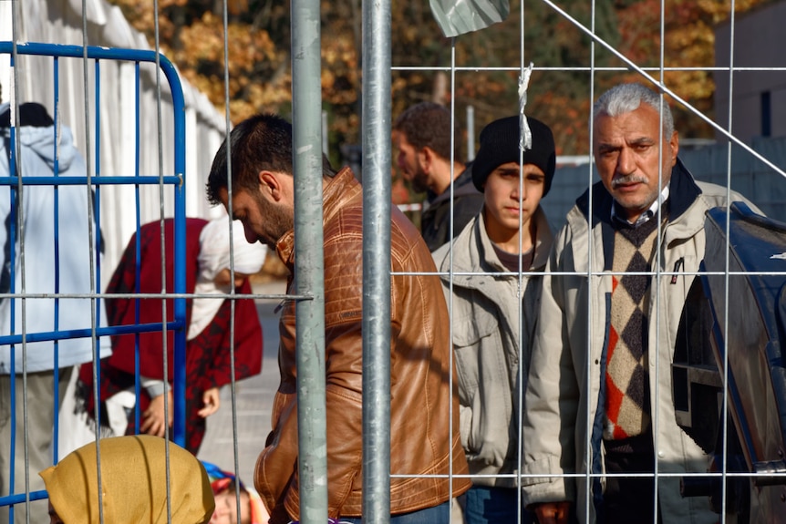 Men stand in line, some looking at the camera through a metal fence.