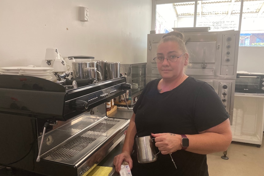 A woman in a black shirt with glasses standing next to a coffee machine and holding a milk jug 