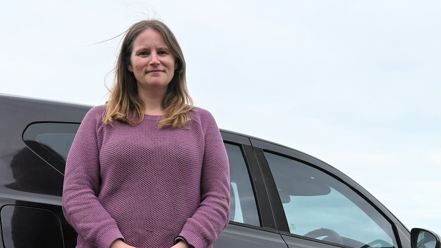 A woman stands in front of a black car in a car park