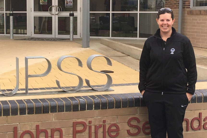 A smiling woman with dark hair and a dark jumper with a logo on it stands in front of a school building next to a J-P-S-S sign
