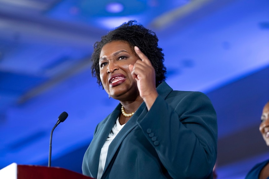 A middle-aged black woman points as she speaks behind a lectern before a blue-tinted backdrop.