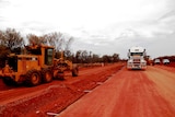 Yellow road work machinery to the left and a road train directly ahead. The red road is dug up in preparation for sealing.