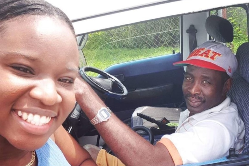 Sam Wallace smiles at the camera as her dad sits inside a white car with a grey cap that says Heat on it
