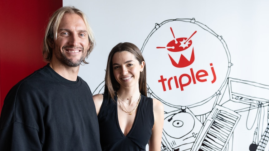 a man and a woman smile at the camera standing in front of a triple j sign 
