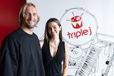 a man and a woman smile at the camera standing in front of a triple j sign 